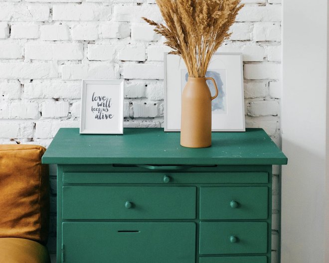 A picture of a green drawer next to a mustard sofa. Over the drawer there's a picture frame with the quote 'Love will keep us alive' and a yellow vase with wheat